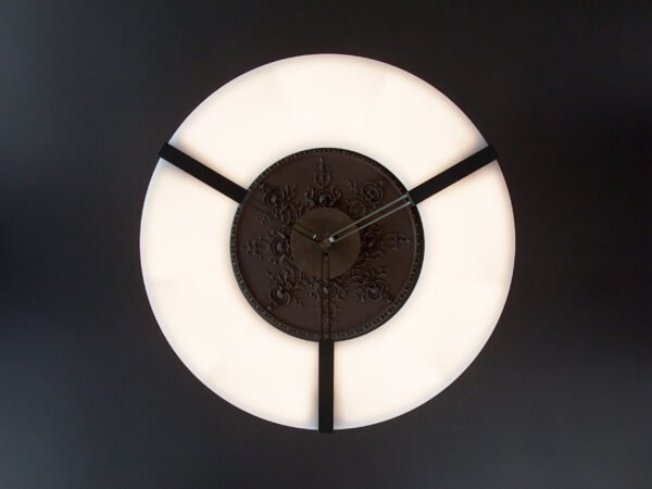 Ozone Light. Le lustre Luxembourg d'Ozone vu de dessous : un large anneau lumineux blanc suspendu par trois bras noirs à une rosace de plafond ornementée.