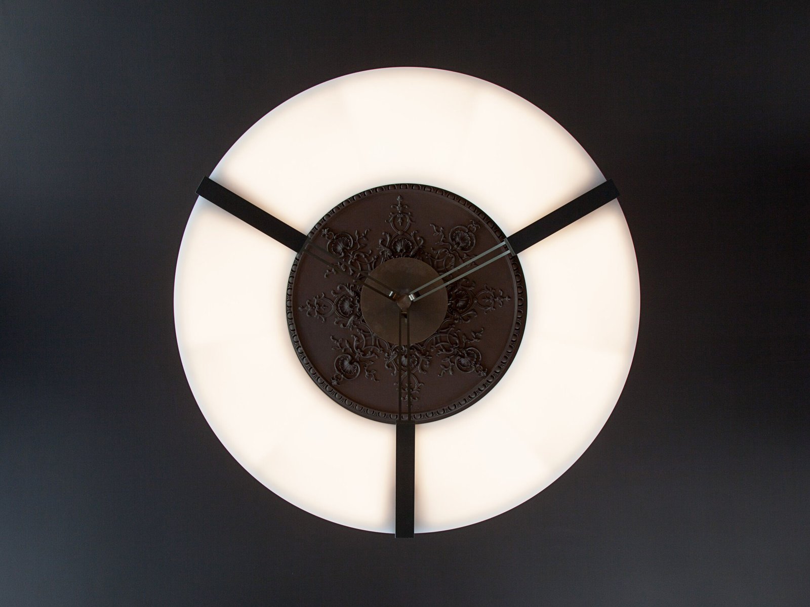 Ozone Light. Le lustre Luxembourg d'Ozone vu de dessous : un large anneau lumineux blanc suspendu par trois bras noirs à une rosace de plafond ornementée.