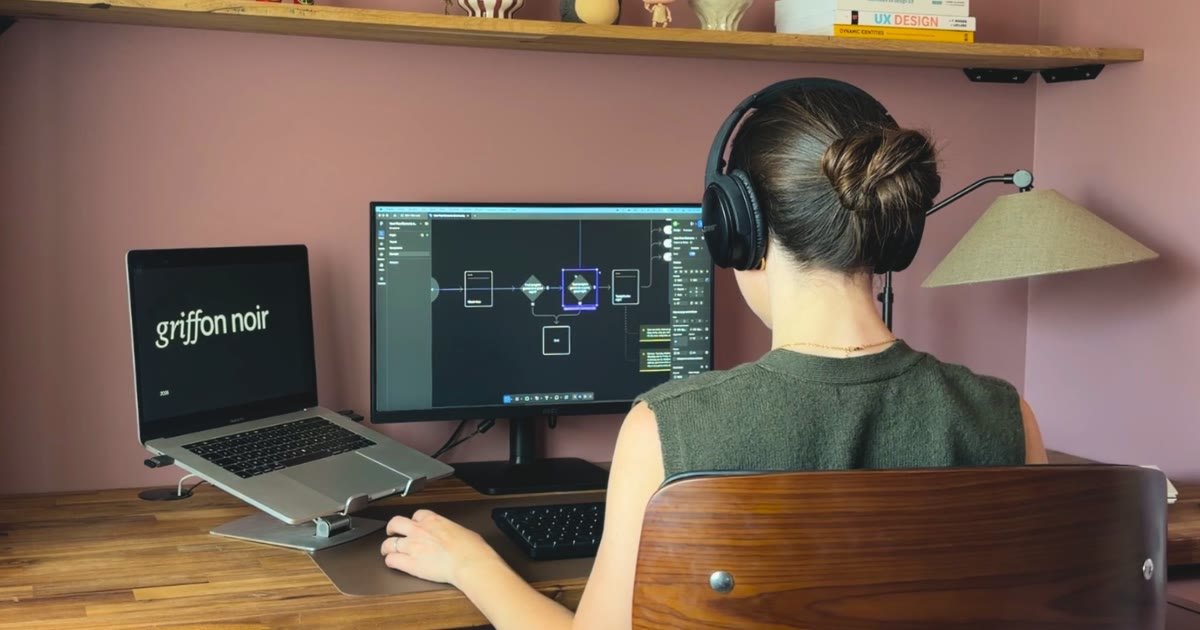 Une designer de dos, casque sur les oreilles, travaille un schéma de parcours sur un grand écran ; à côté, un ordinateur portable affiche le logo Griffon Noir.